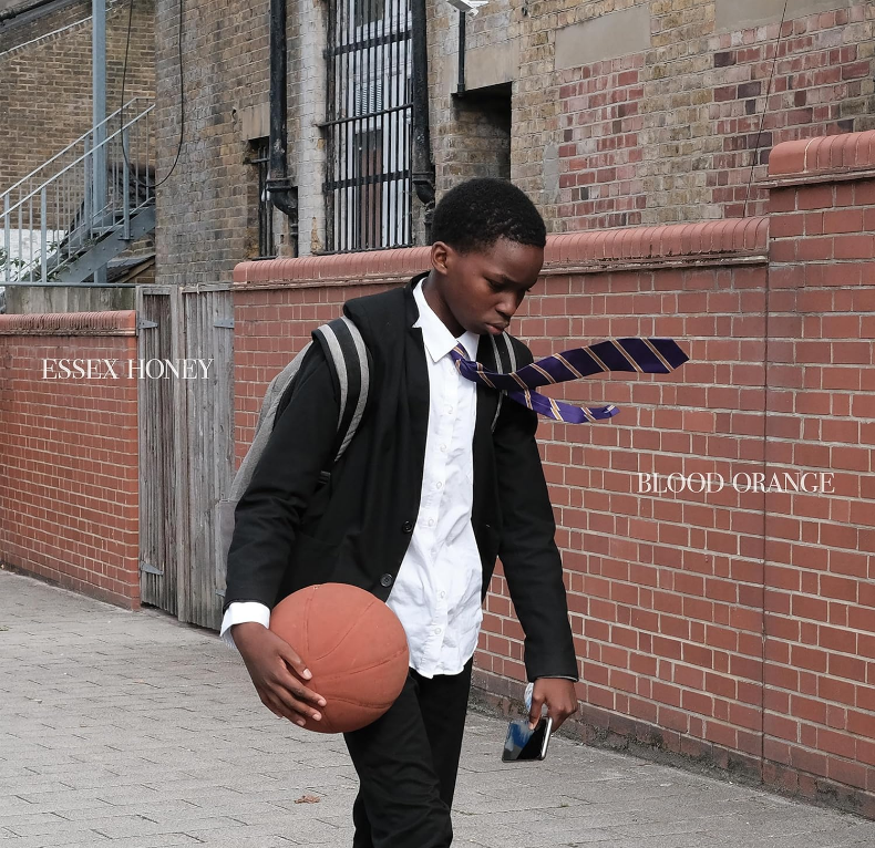 Adolescent Black Boy In a Black Suit, White Shirt, Dark and Tie, Wearing A Backback, And Holding A Basketball In His Right Hand, Walking In Front Of A Brick Wall.